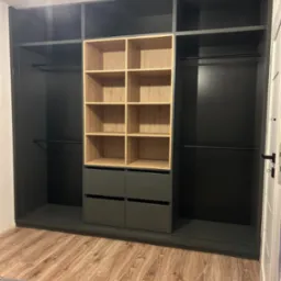 Szara szafa wnękowa z drewnianymi półkami i szufladami, widoczna z perspektywy podłogi, z fragmentem łóżka w dolnej części kadru.