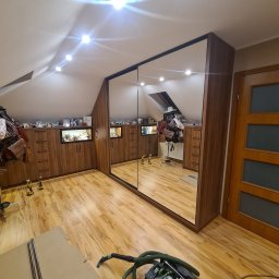 Rafał Bojek - Montaż szafy wnękowej z lustrzanymi drzwiami przesuwnymi w pokoju na poddaszu, widoczne narzędzia i odkurzacz przemysłowy, drewniane wykończenie mebli.