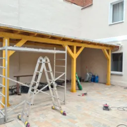 Żółta konstrukcja z drewna, prawdopodobnie w trakcie budowy, widoczna drabina, narzędzia i materiały budowlane na brukowanym podwórku.