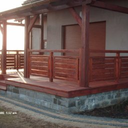 Rafał Bojek - Zadaszony taras drewniany z niską balustradą z poziomych desek, wsparty na fundamencie z szarych bloczków betonowych, z widocznym fragmentem rynny i rolet zewnętrznych w tle.