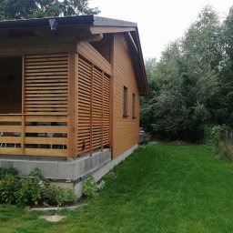 Rafał Bojek - Drewniany domek letniskowy z zadaszonym tarasem, wykończony jasnym drewnem, widok z boku na zadbany trawnik i drzewa w tle.