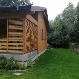 Drewniany domek letniskowy z zadaszonym tarasem, wykończony jasnym drewnem, widok z boku na zadbany trawnik i drzewa w tle.