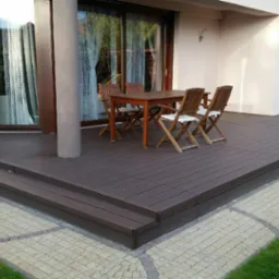 Brunatny taras z kompozytu drewna, z niskimi schodami, otoczony kostką brukową i trawnikiem, z drewnianym stołem i krzesłami na wierzchu, przylegający do jasnego budynku z dużymi oknami.