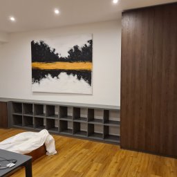 Rafał Bojek - Wnętrze pokoju z zabudową meblową: wysoka szafa z ciemnego drewna i szara, modułowa półka na ścianie, nad nią abstrakcyjny obraz. Podłoga laminowana, w tle widoczny stół z laptopem.