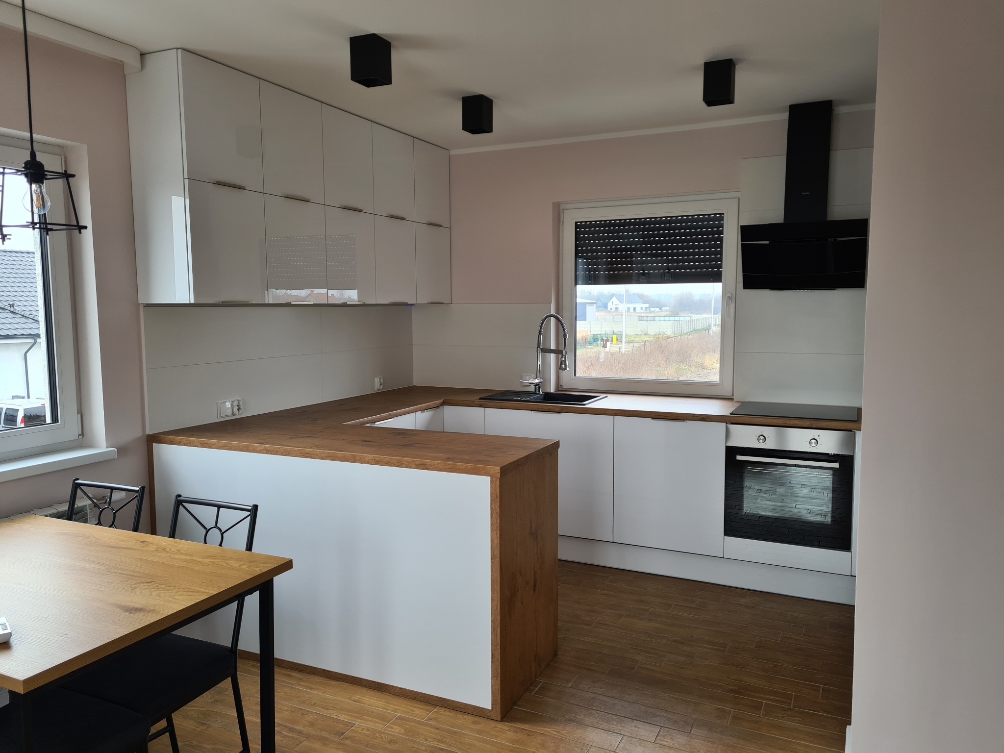 Nowoczesna kuchnia z białymi szafkami i drewnianym blatem, czarne lampy sufitowe, drewniany stół z czarnymi krzesłami, widok na zewnątrz przez okno.