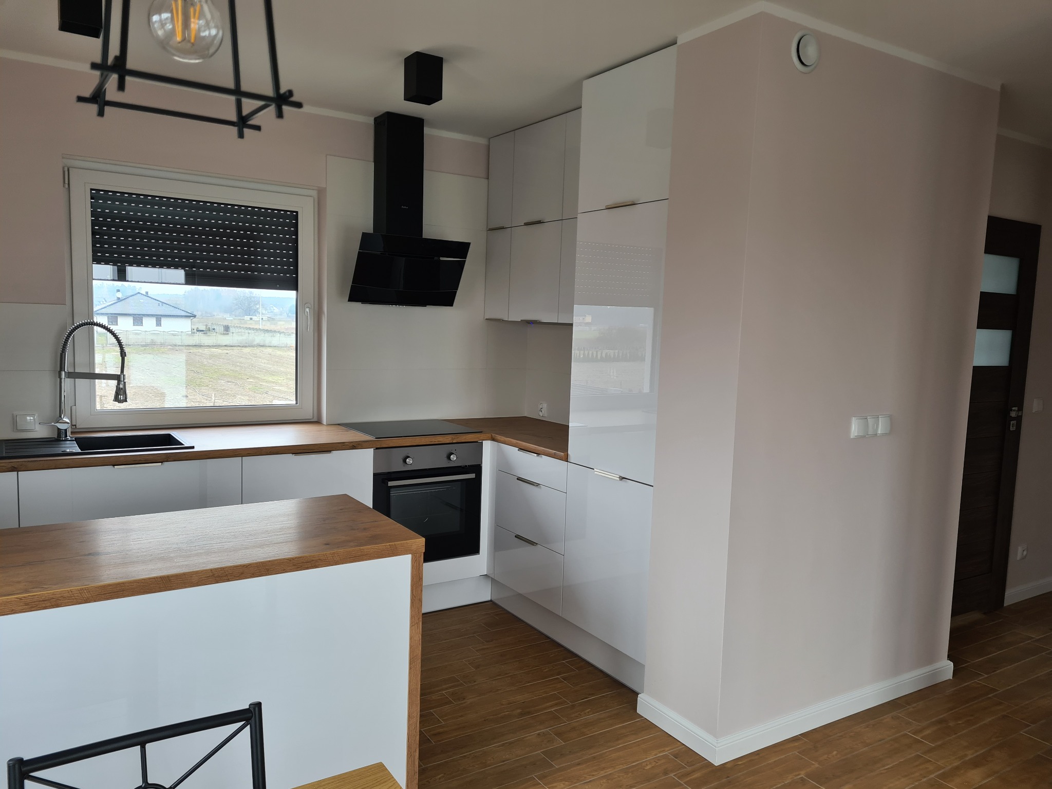 Nowoczesna kuchnia z białymi, lakierowanymi szafkami, drewnianym blatem, czarnym okapem i widokiem na dom zza okna.