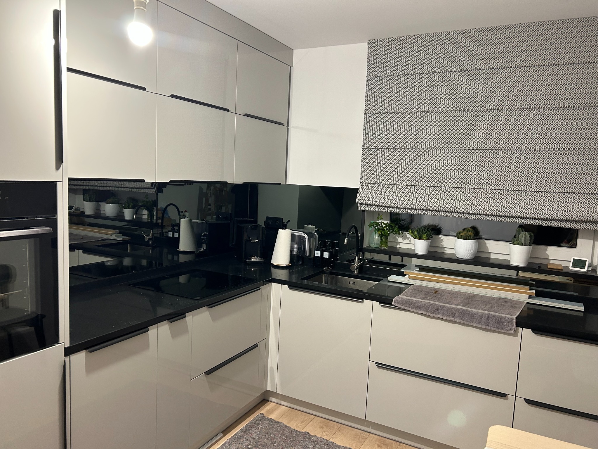 Nowoczesna kuchnia w odcieniach szarości z czarnym blatem i minimalistycznymi uchwytami, uzupełniona roletą rzymską w geometryczny wzór i doniczkami z sukulentami na parapecie.