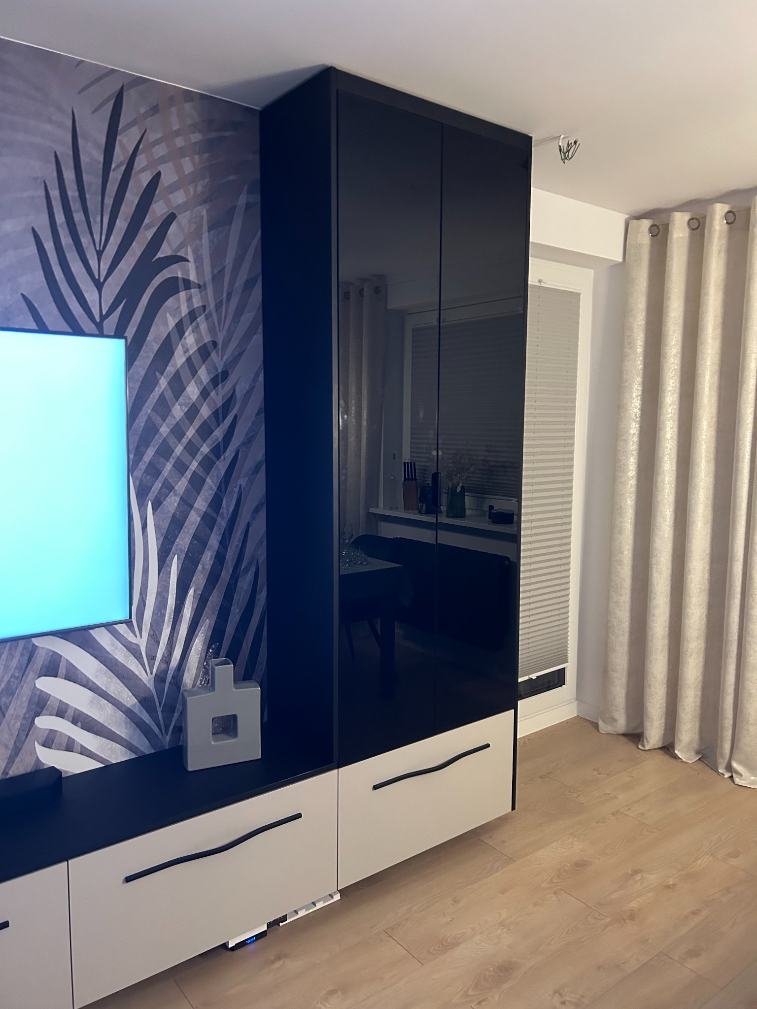 Częściowy widok nowoczesnej szafy wnękowej z czarnymi, lakierowanymi frontami i białymi szufladami, dopasowanej do ściany z tapetą w motywy roślinne, uzupełnionej o dekoracyjny wazon.