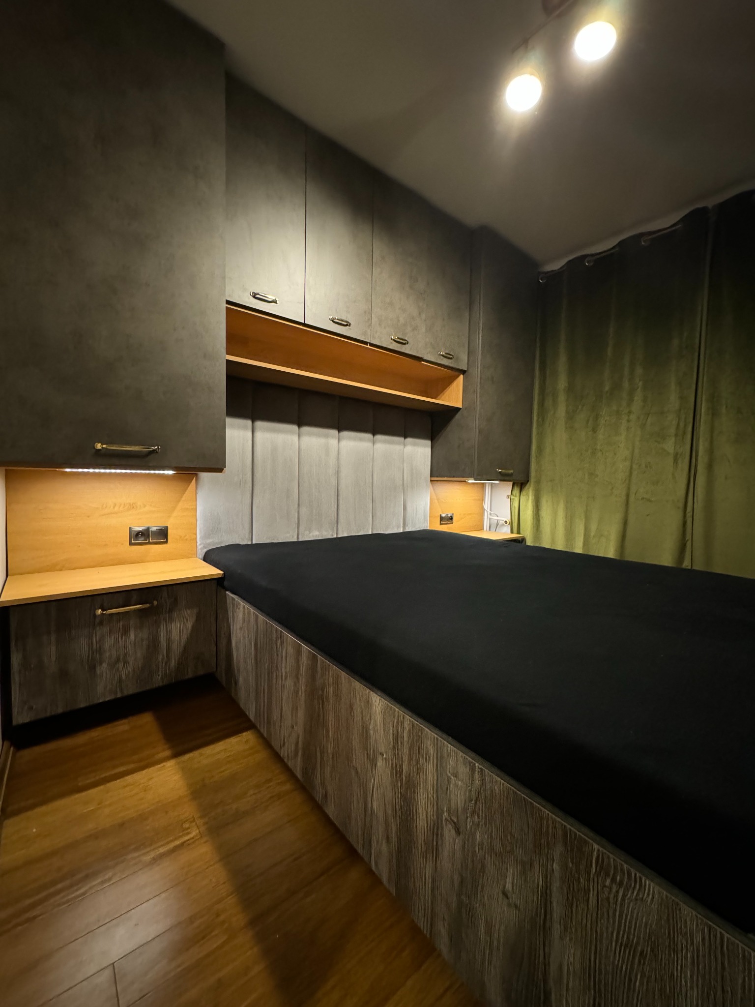 Nowoczesna sypialnia z łóżkiem w zabudowie, szafkami nocnymi i szafkami wiszącymi w odcieniach szarości i drewna, z oświetleniem LED i ciemną pościelą.