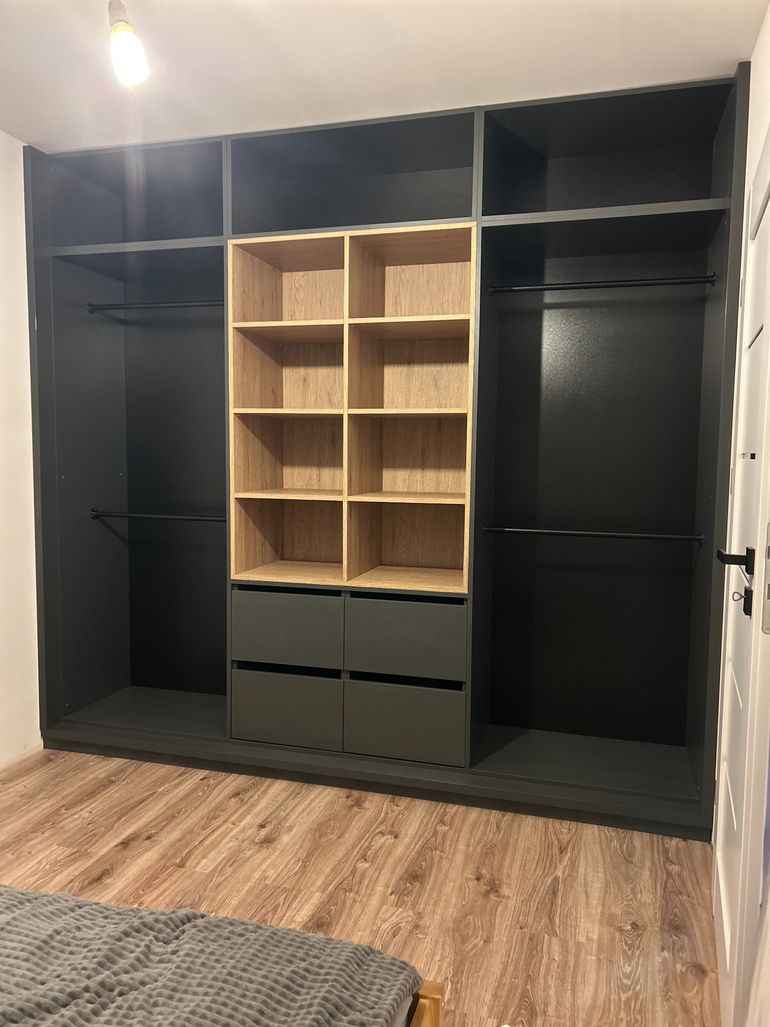 Szara szafa wnękowa z drewnianymi półkami i szufladami, widoczna z perspektywy podłogi, z fragmentem łóżka w dolnej części kadru.