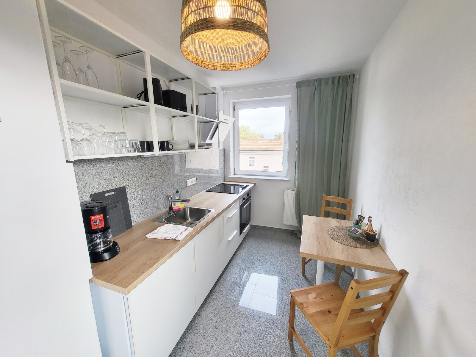 Jasna kuchnia z białymi szafkami, drewnianym blatem, okrągłym żyrandolem i drewnianym stołem z dwoma krzesłami, widok na zewnątrz przez okno.