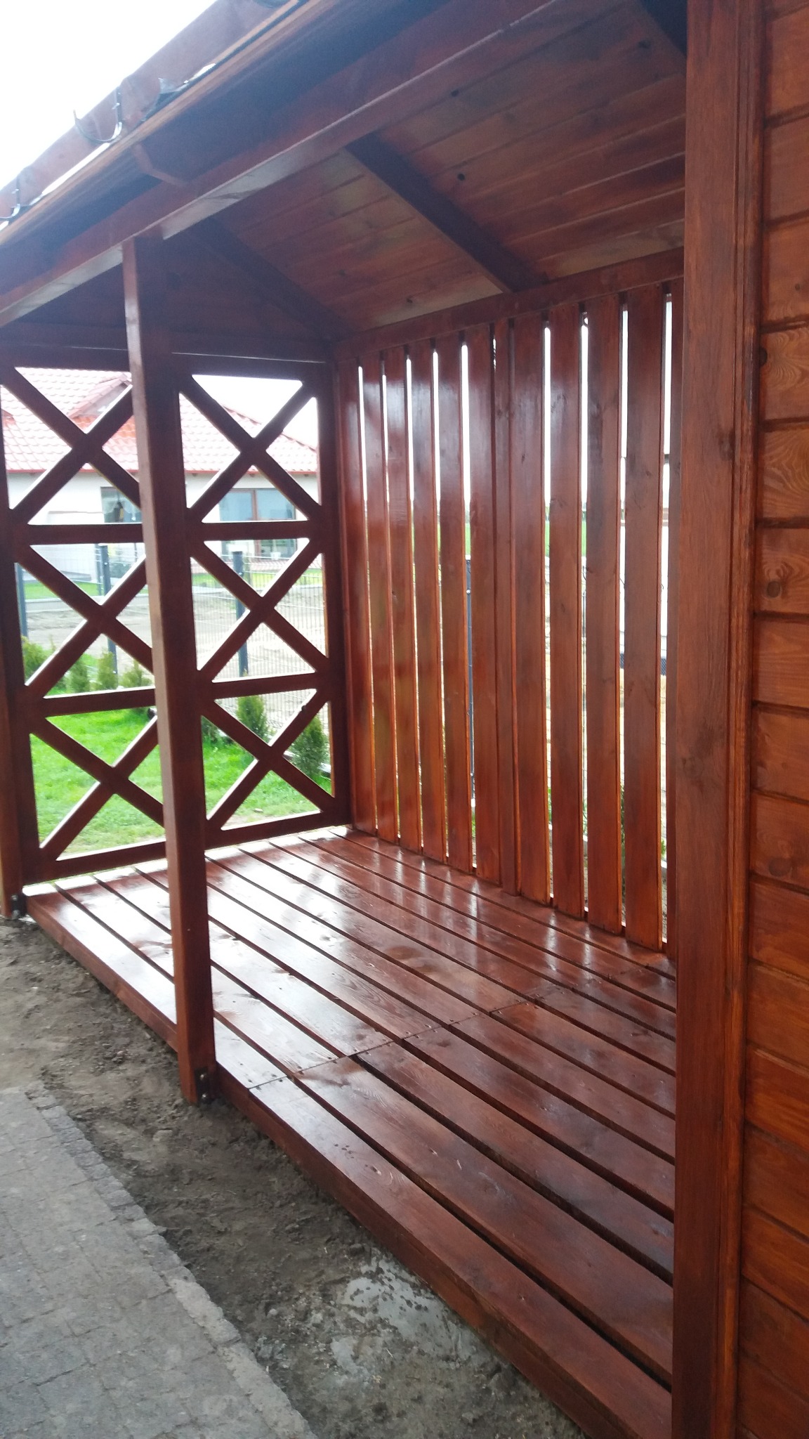 Nowa, drewniana altana z dachem, pomalowana na brązowo, z widoczną konstrukcją kratową i pionowymi deskami, świeżo po impregnacji, z widokiem na trawnik i dom w tle.