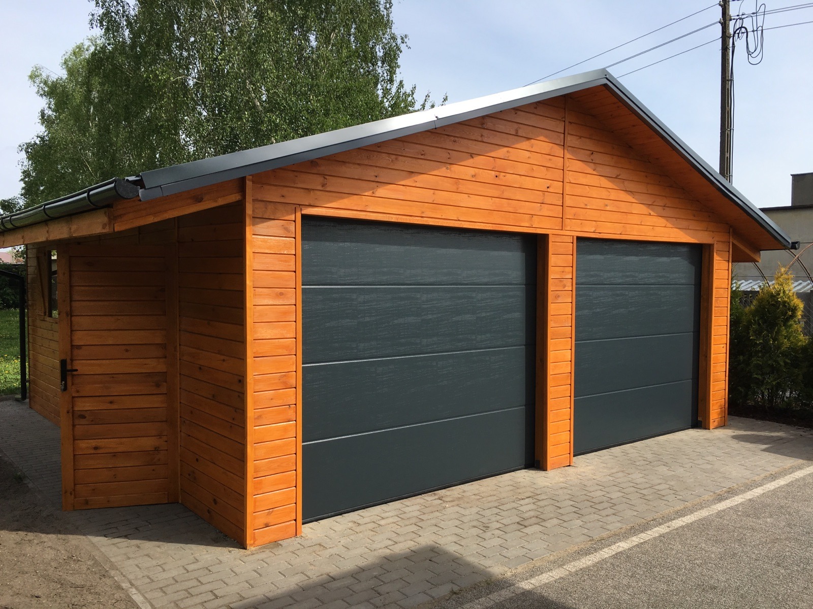 Drewniany garaż dwustanowiskowy z szarymi segmentowymi bramami, widoczny z zewnątrz, z dodatkowym pomieszczeniem z boku.
