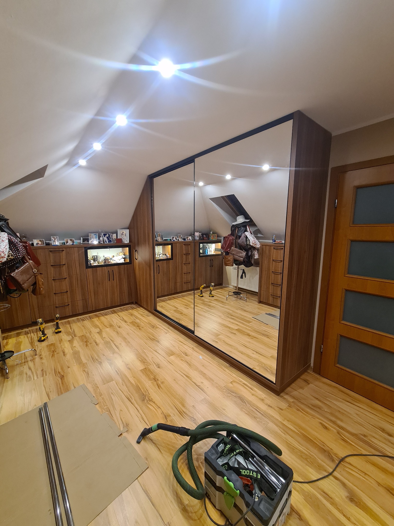 Montaż szafy wnękowej z lustrzanymi drzwiami przesuwnymi w pokoju na poddaszu, widoczne narzędzia i odkurzacz przemysłowy, drewniane wykończenie mebli.
