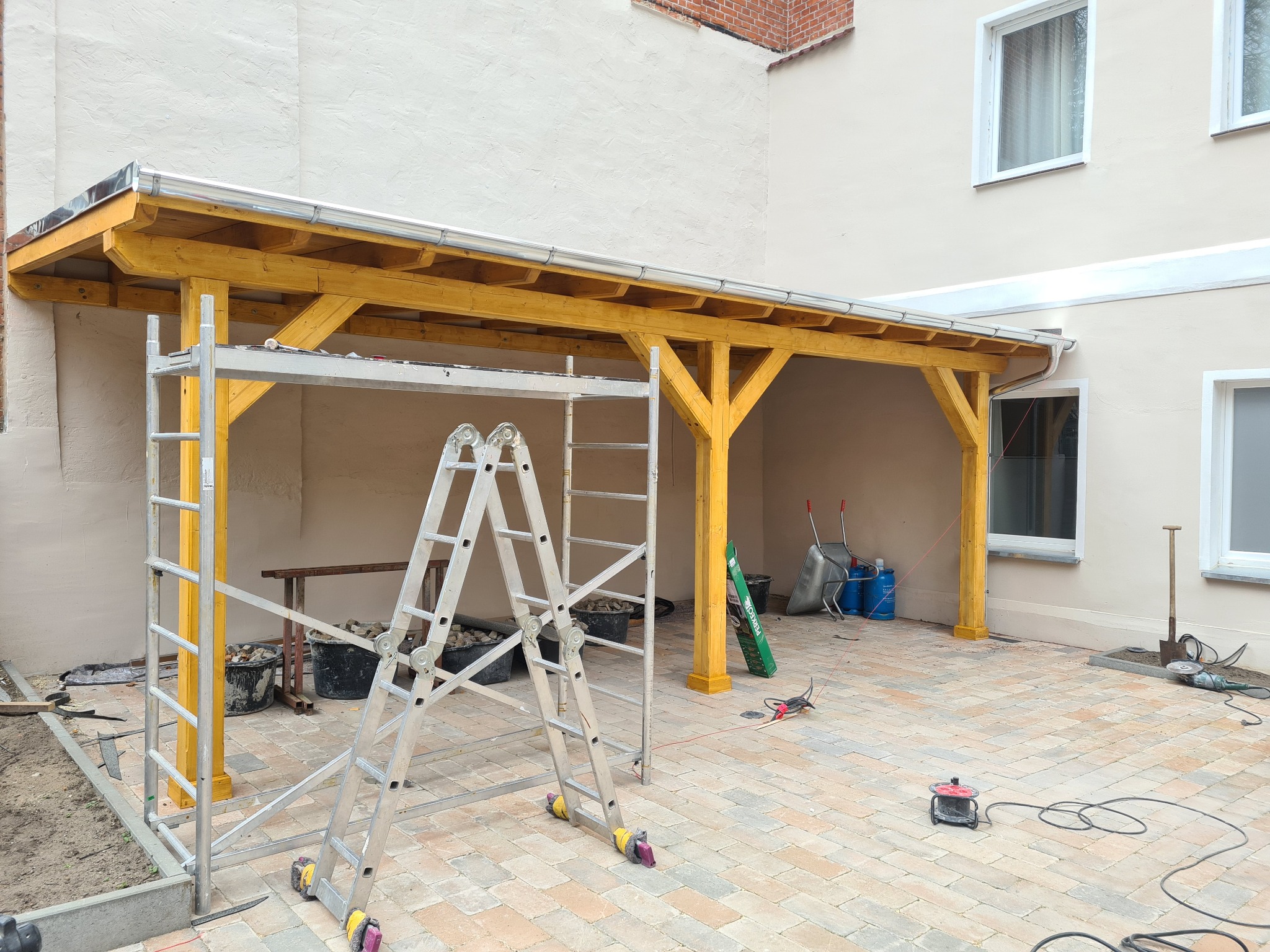 Żółta konstrukcja z drewna, prawdopodobnie w trakcie budowy, widoczna drabina, narzędzia i materiały budowlane na brukowanym podwórku.