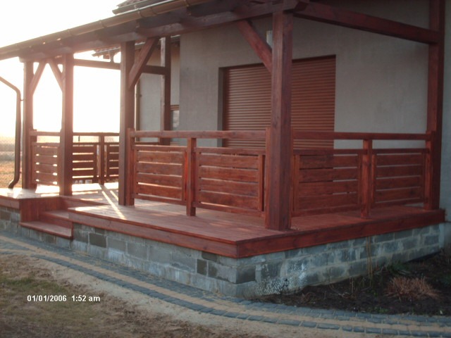 Zadaszony taras drewniany z niską balustradą z poziomych desek, wsparty na fundamencie z szarych bloczków betonowych, z widocznym fragmentem rynny i rolet zewnętrznych w tle.