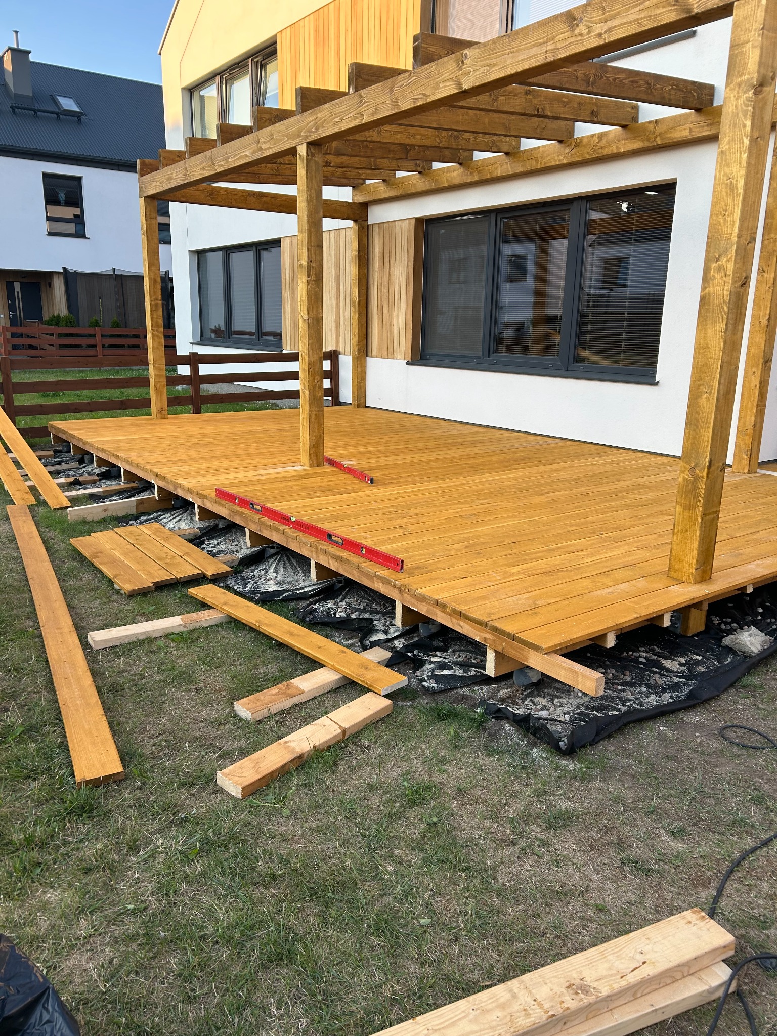 W trakcie budowy drewnianego tarasu z pergolą przy nowoczesnym domu; widoczne deski, poziomica, czarna folia izolacyjna i elementy konstrukcyjne pergoli.