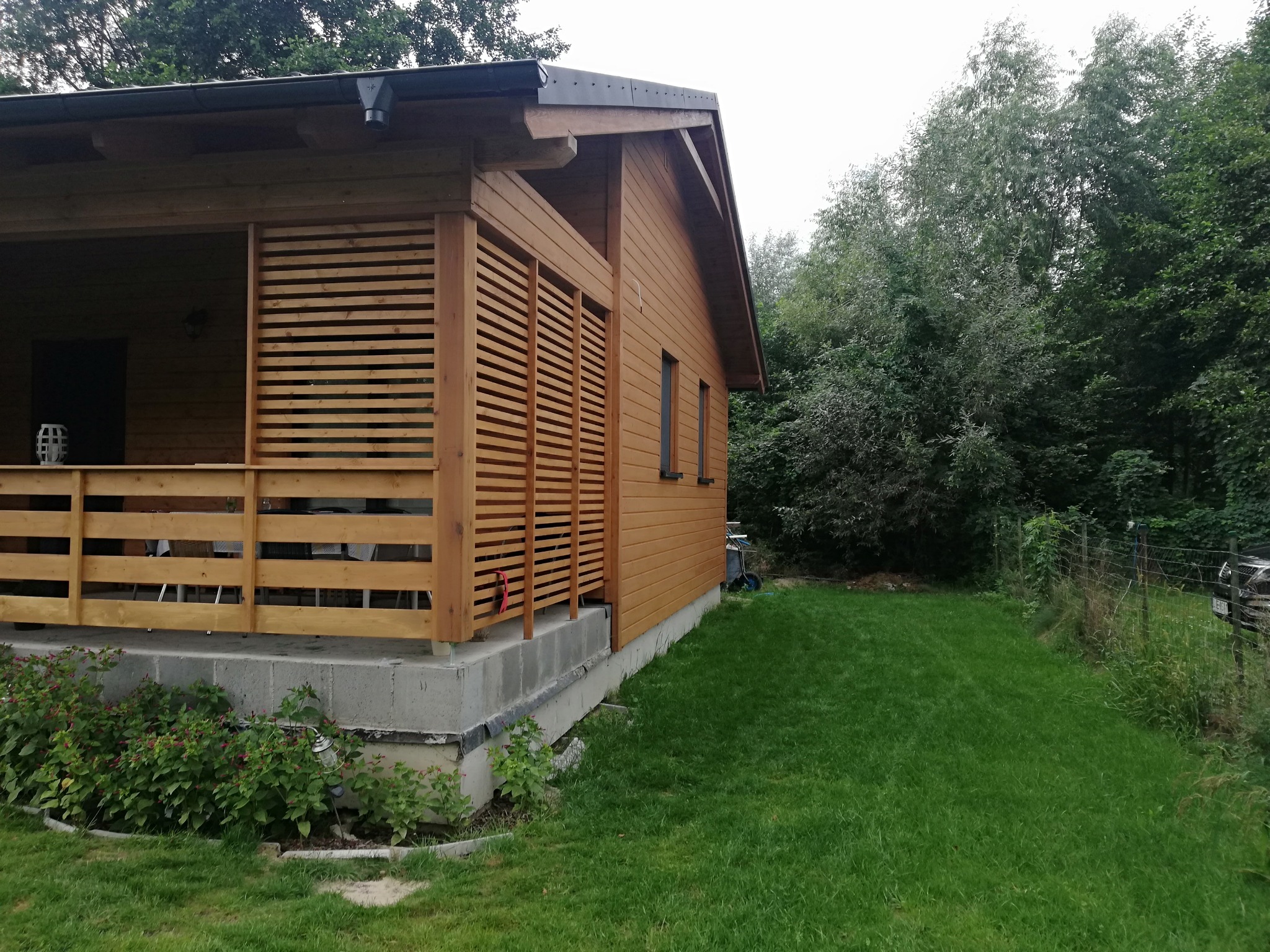 Drewniany domek letniskowy z zadaszonym tarasem, wykończony jasnym drewnem, widok z boku na zadbany trawnik i drzewa w tle.