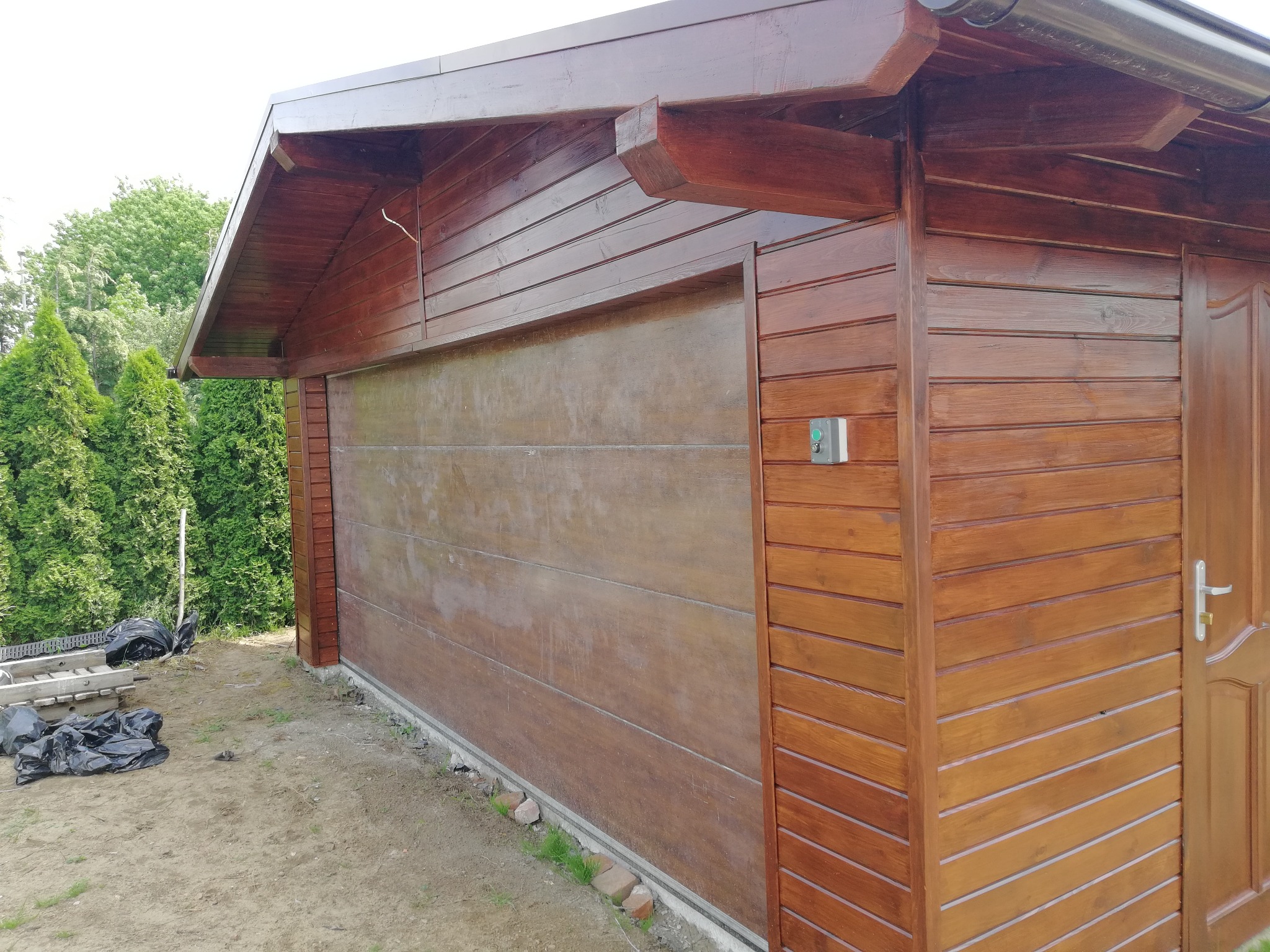 Drewniany garaż z automatyczną bramą segmentową i drzwiami wejściowymi, wykończony brązową bejcą, widok z zewnątrz, obok trawnik i drabina.