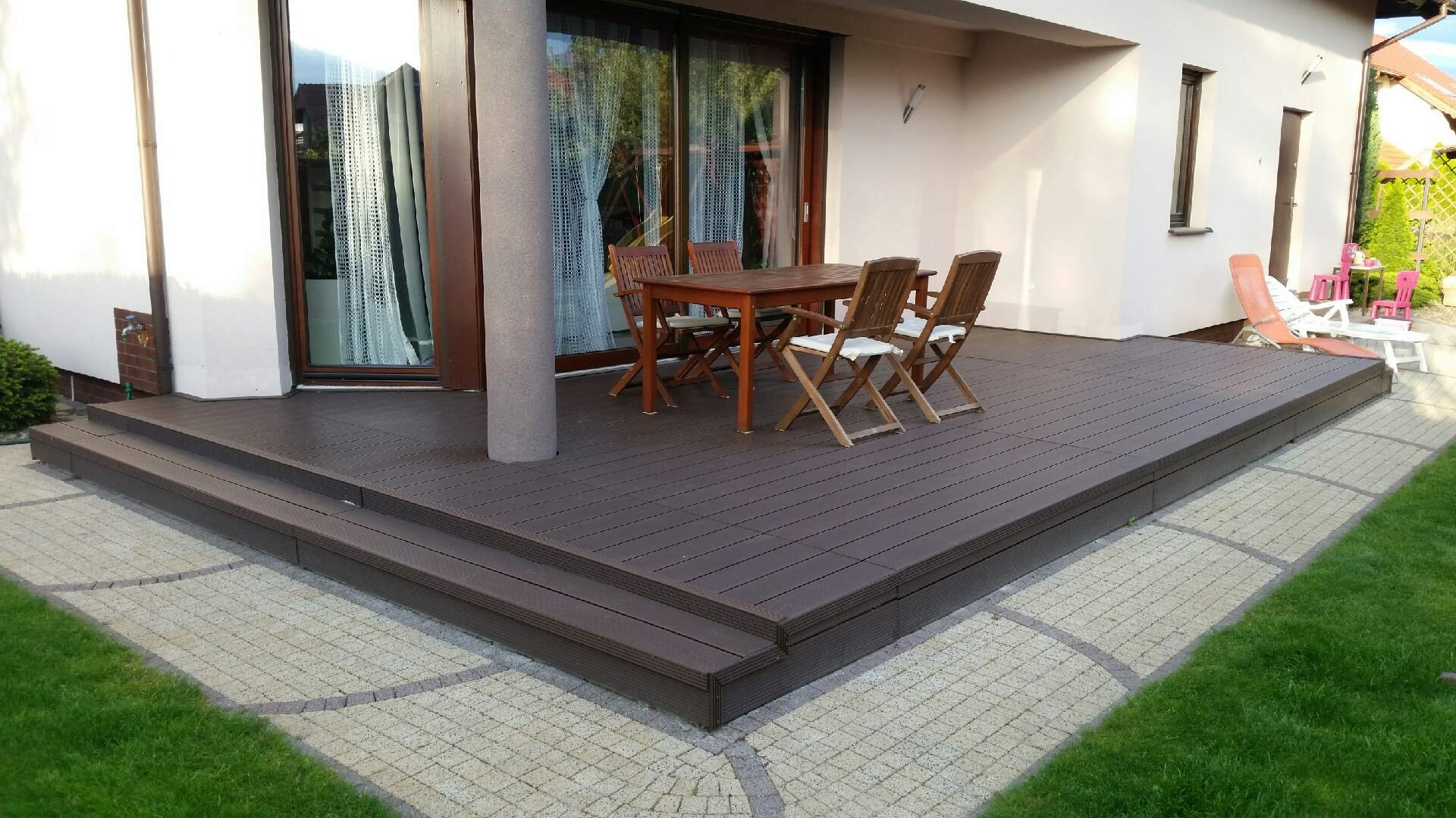 Brunatny taras z kompozytu drewna, z niskimi schodami, otoczony kostką brukową i trawnikiem, z drewnianym stołem i krzesłami na wierzchu, przylegający do jasnego budynku z dużymi oknami.