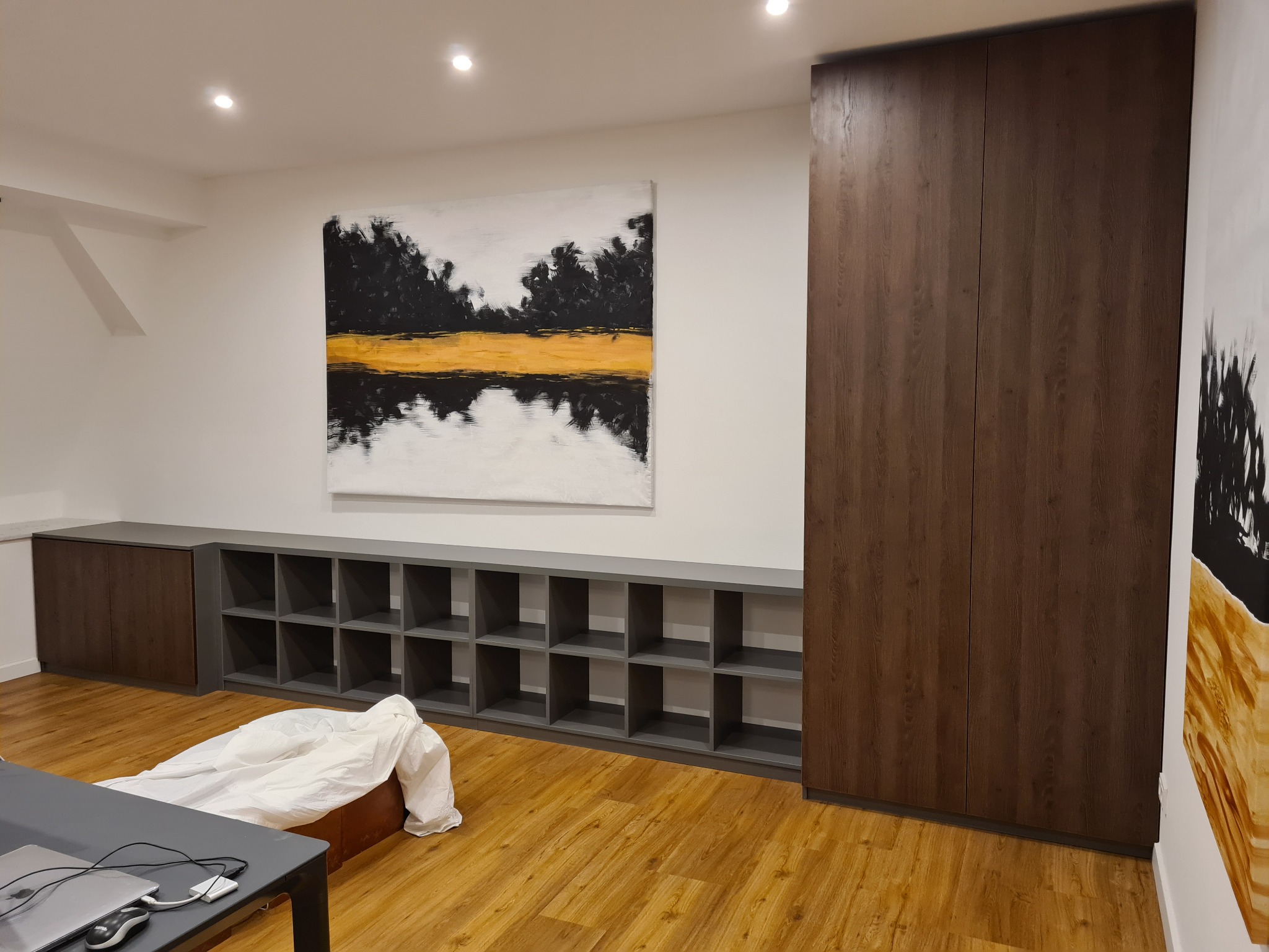 Wnętrze pokoju z zabudową meblową: wysoka szafa z ciemnego drewna i szara, modułowa półka na ścianie, nad nią abstrakcyjny obraz. Podłoga laminowana, w tle widoczny stół z laptopem.