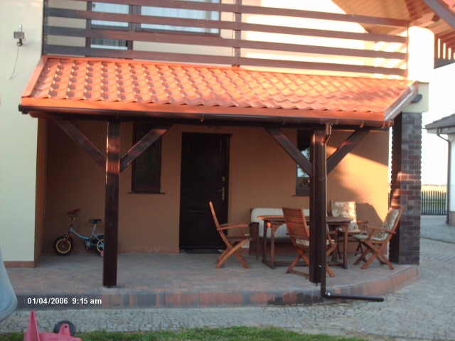 Zadaszony taras z brązową konstrukcją drewnianą i czerwoną dachówką, widoczny komplet mebli ogrodowych i dziecięcy rowerek.