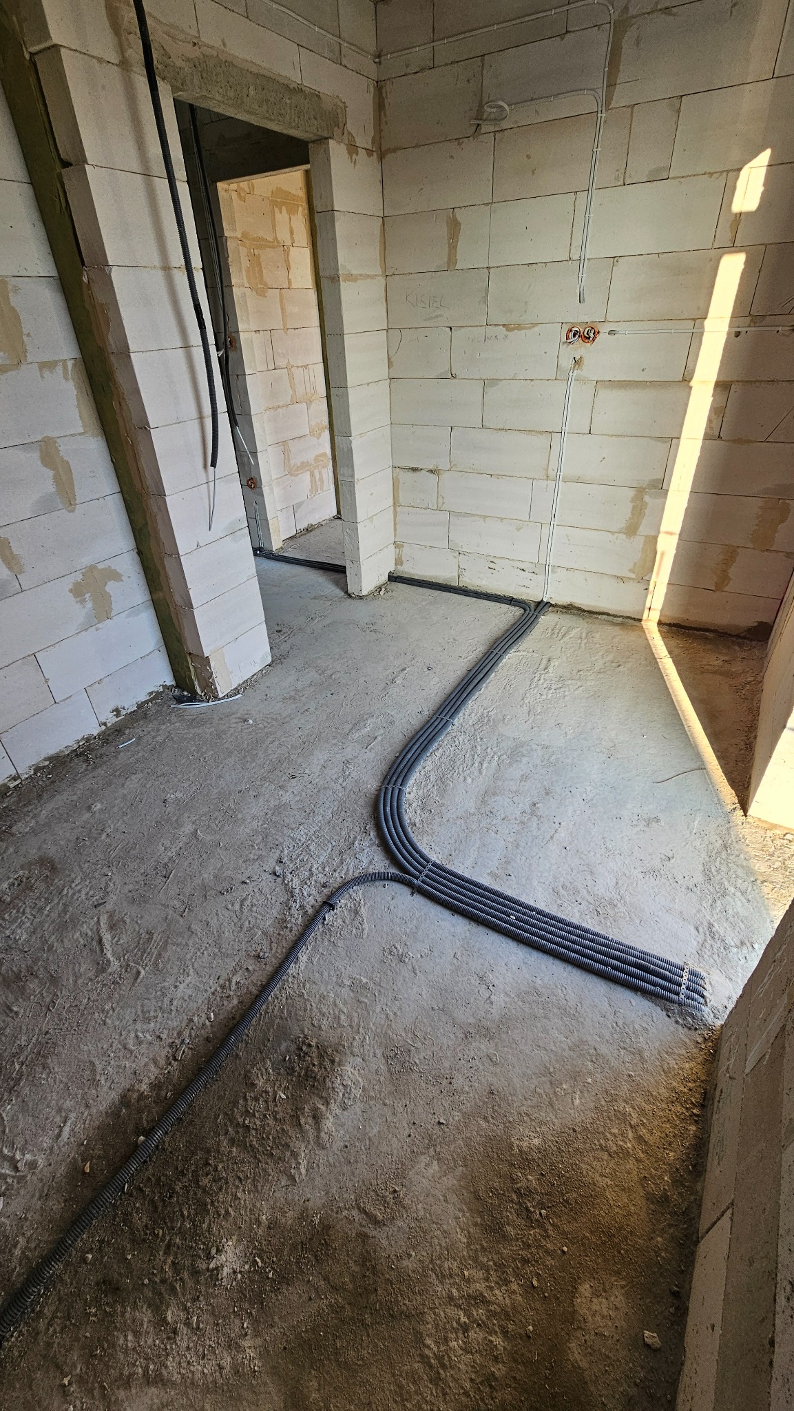 Wnętrze surowego budynku w trakcie prac elektrycznych: kable w peszlach prowadzone po podłodze, puszki elektryczne zamontowane w ścianie z betonu komórkowego.