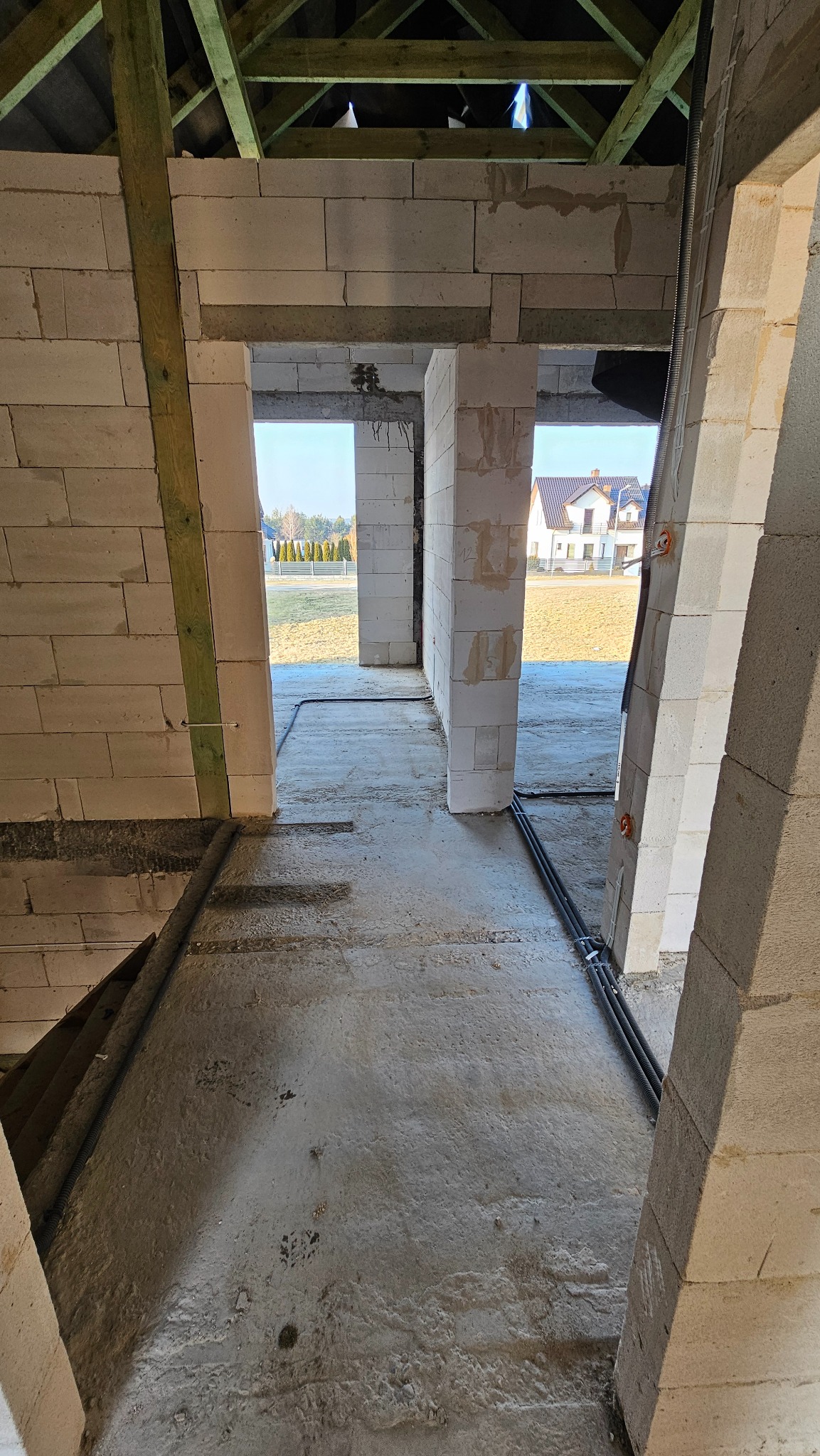 Wnętrze surowego budynku w trakcie budowy z widocznymi przewodami elektrycznymi w peszlach ułożonymi na posadzce, bloczki betonowe ścian, drewniana konstrukcja dachu, w tle dom jednorodzinny.