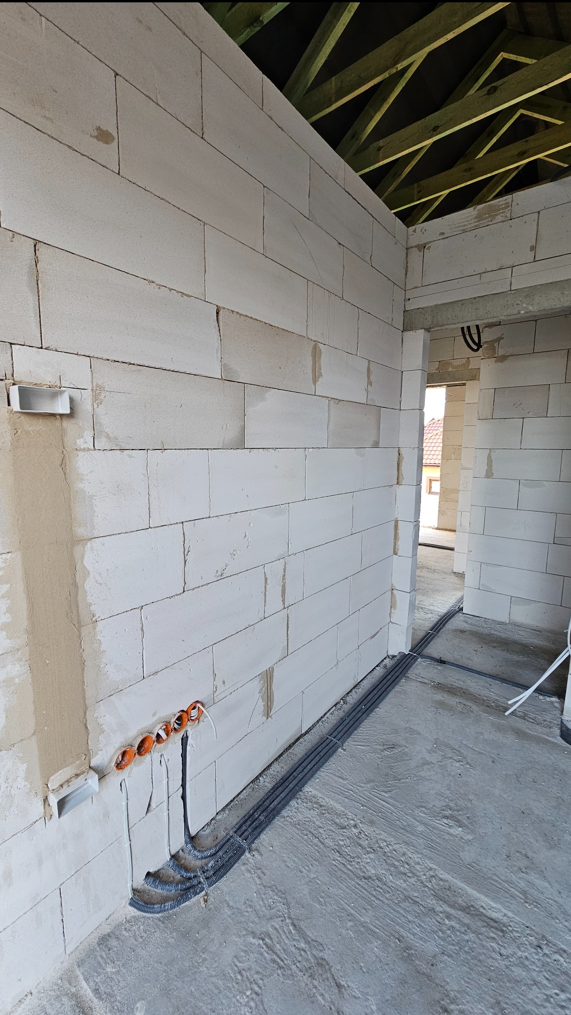 Wewnętrzne ściany z betonu komórkowego w surowym stanie z widocznymi puszkami elektrycznymi i peszlami ułożonymi wzdłuż podłogi, widoczne elementy więźby dachowej.