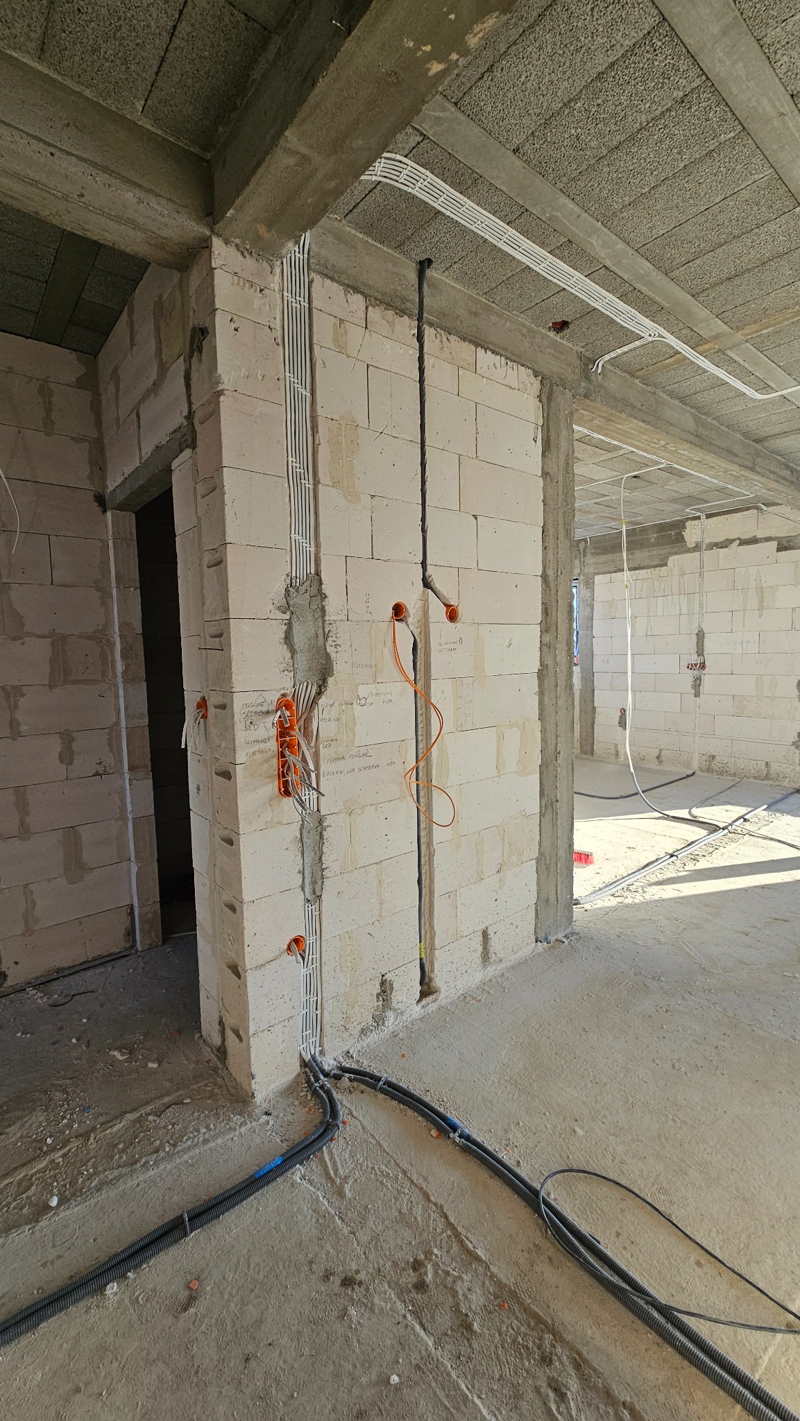 Surowy stan budynku w trakcie prac elektrycznych: widoczne kable w korytach i peszlach, pomarańczowa puszka podtynkowa zalana gipsem, przewody poprowadzone w bruzdach w ścianie z betonu komórkowego.