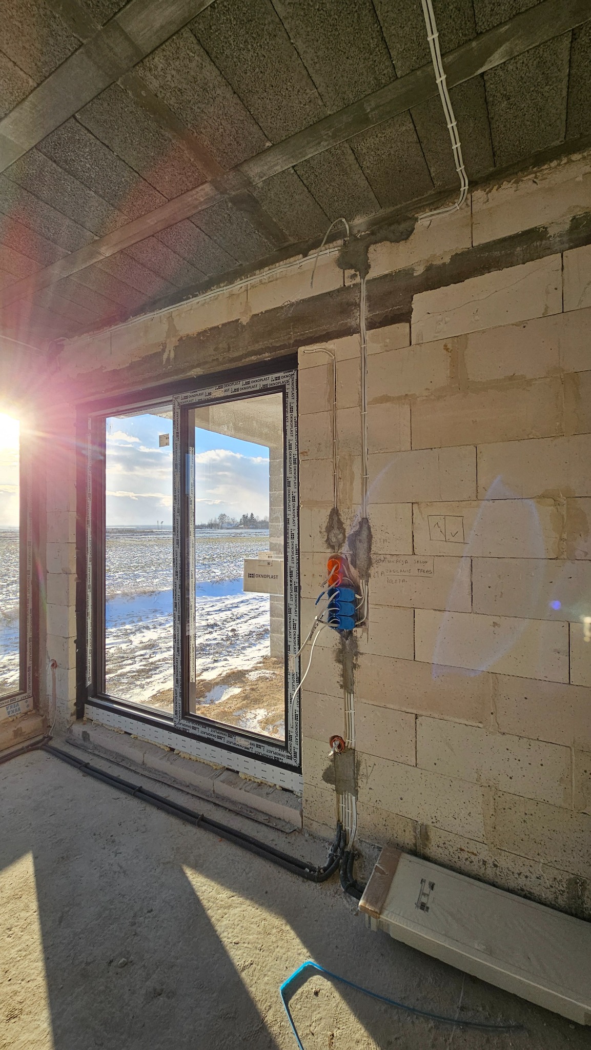 Instalacja elektryczna w surowym stanie budynku, widoczne przewody prowadzone po ścianie z betonu komórkowego, puszki elektryczne w kolorze pomarańczowym i niebieskim, okno z widokiem na zaśnieżone...