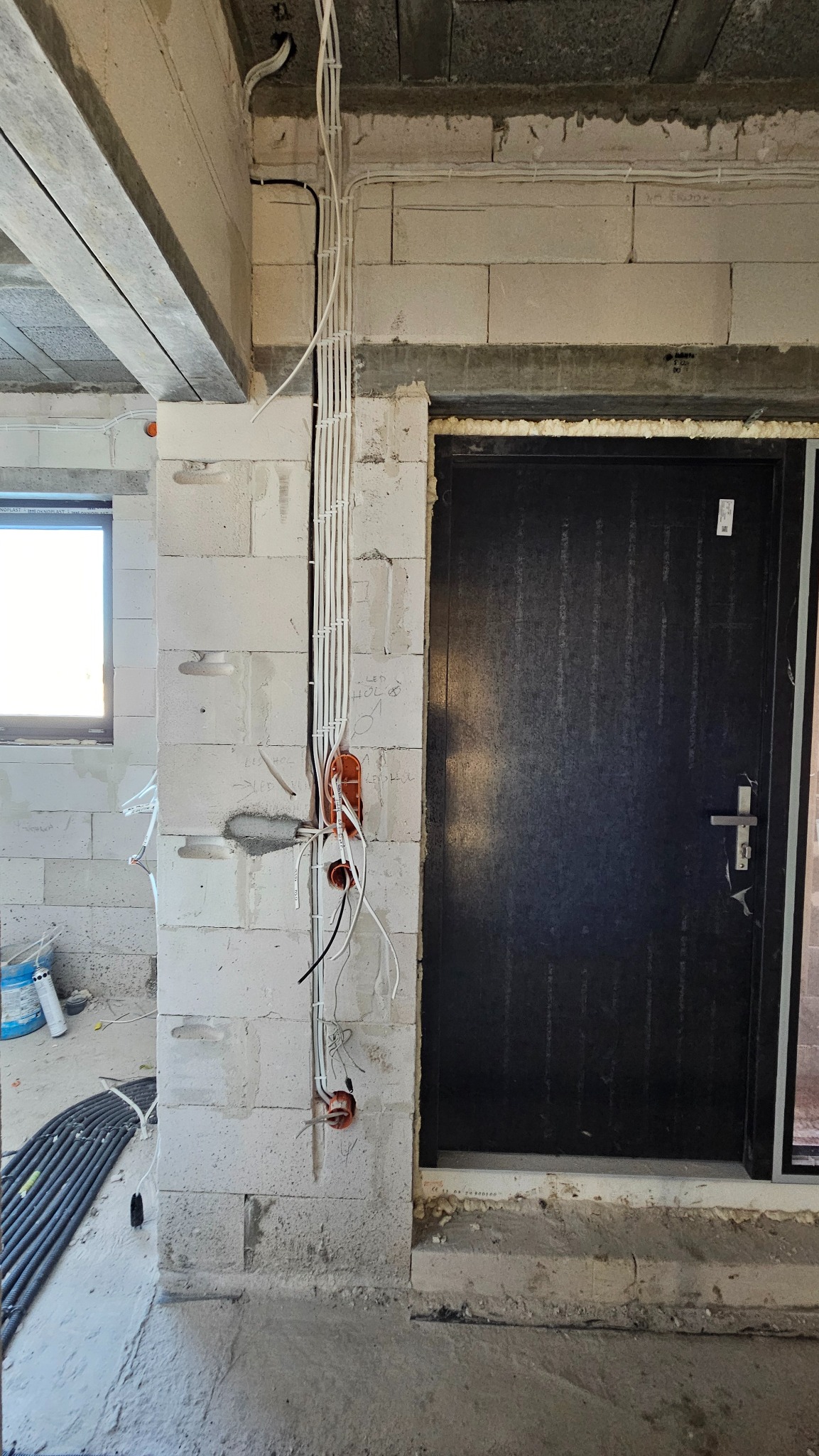 Surowe wnętrze w trakcie budowy, widoczne przewody elektryczne prowadzone po ścianie z bloczków, puszki instalacyjne, czarne drzwi wejściowe, betonowa podłoga i sufit.