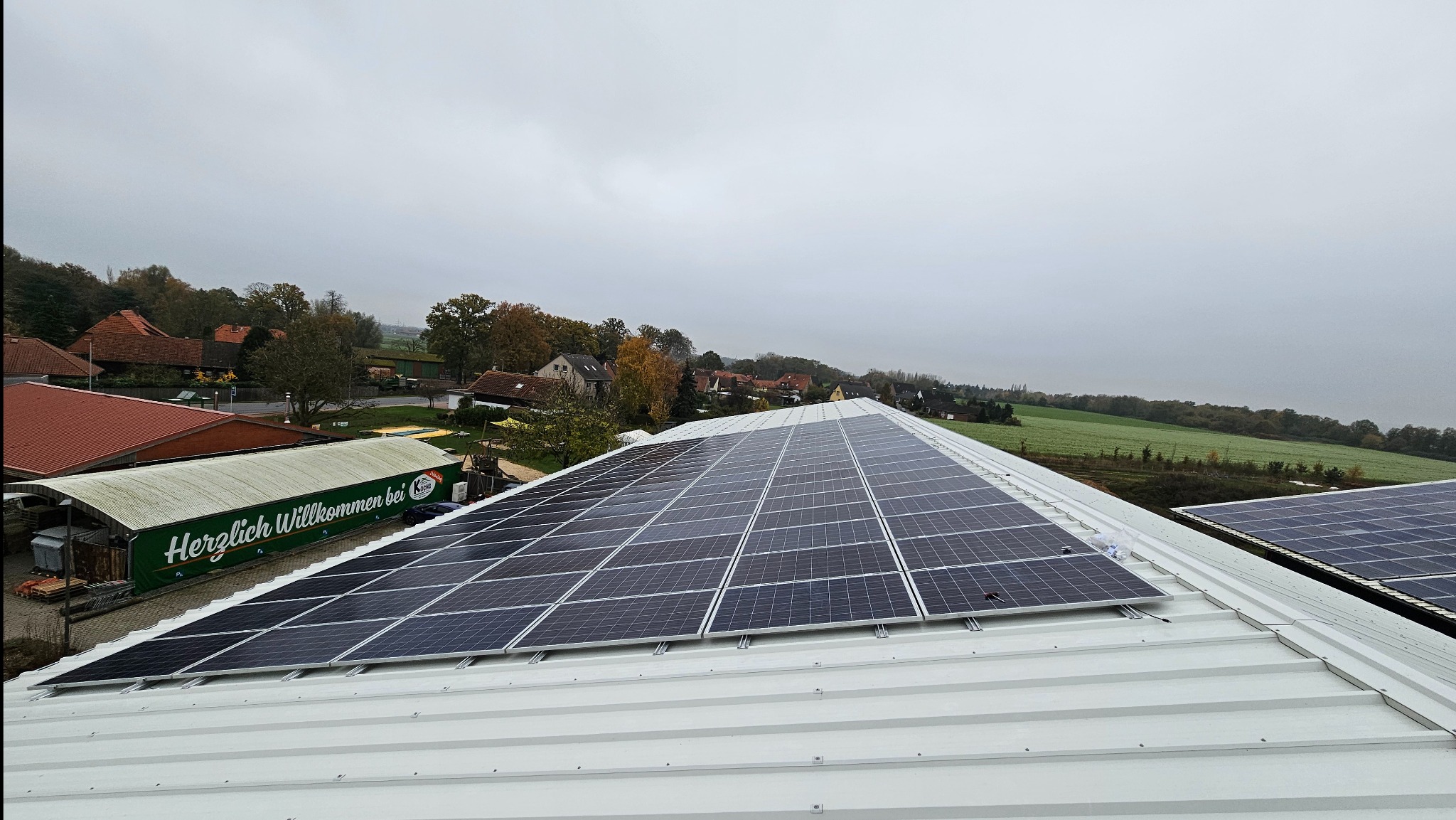 Widok z góry na panele fotowoltaiczne zamontowane na białym blaszanym dachu budynku gospodarczego, w tle zabudowania wiejskie i pochmurne niebo.
