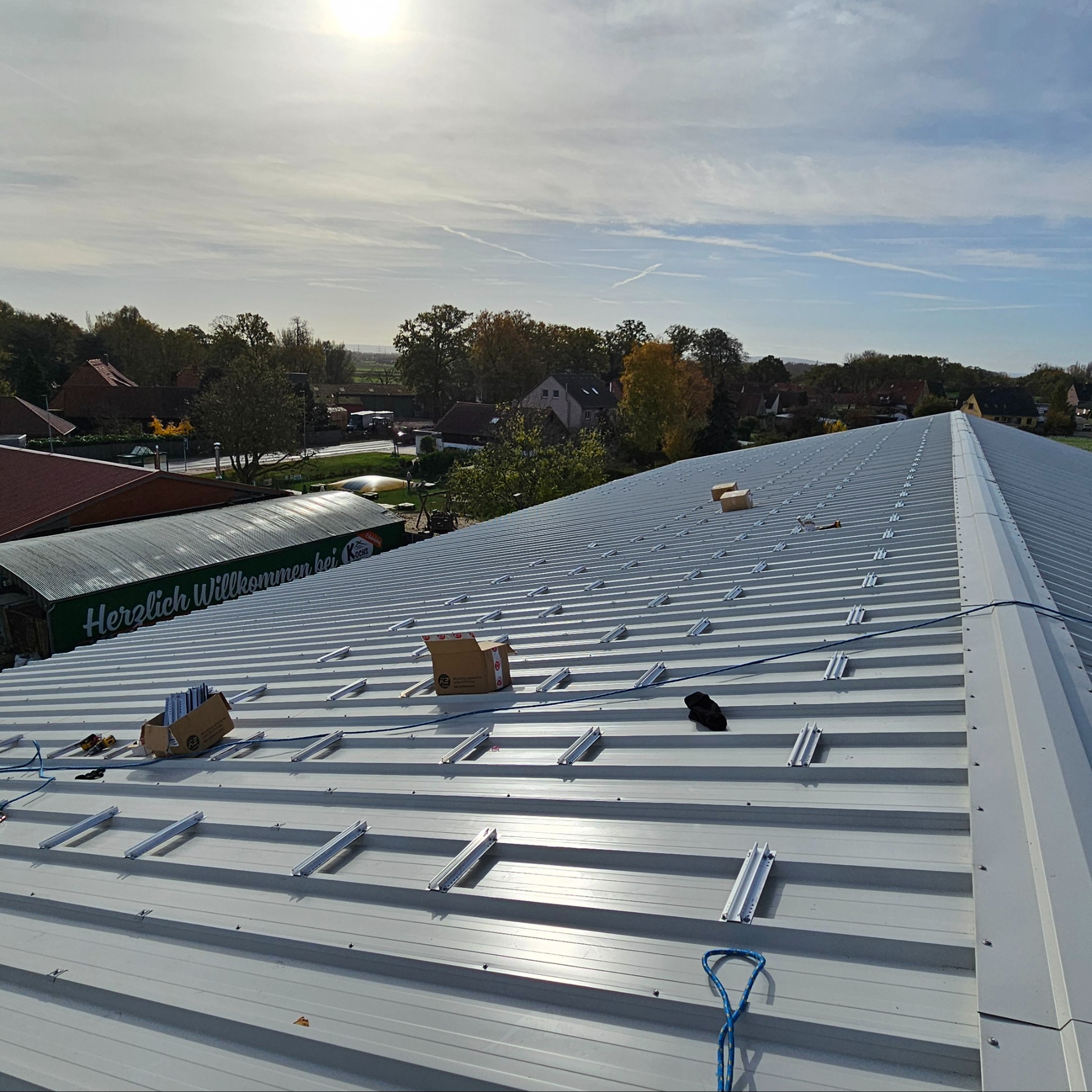 Montaż systemu mocowania paneli fotowoltaicznych na blaszanym dachu z widocznymi elementami konstrukcyjnymi, narzędziami i pudełkami z akcesoriami, w tle budynki i drzewa.