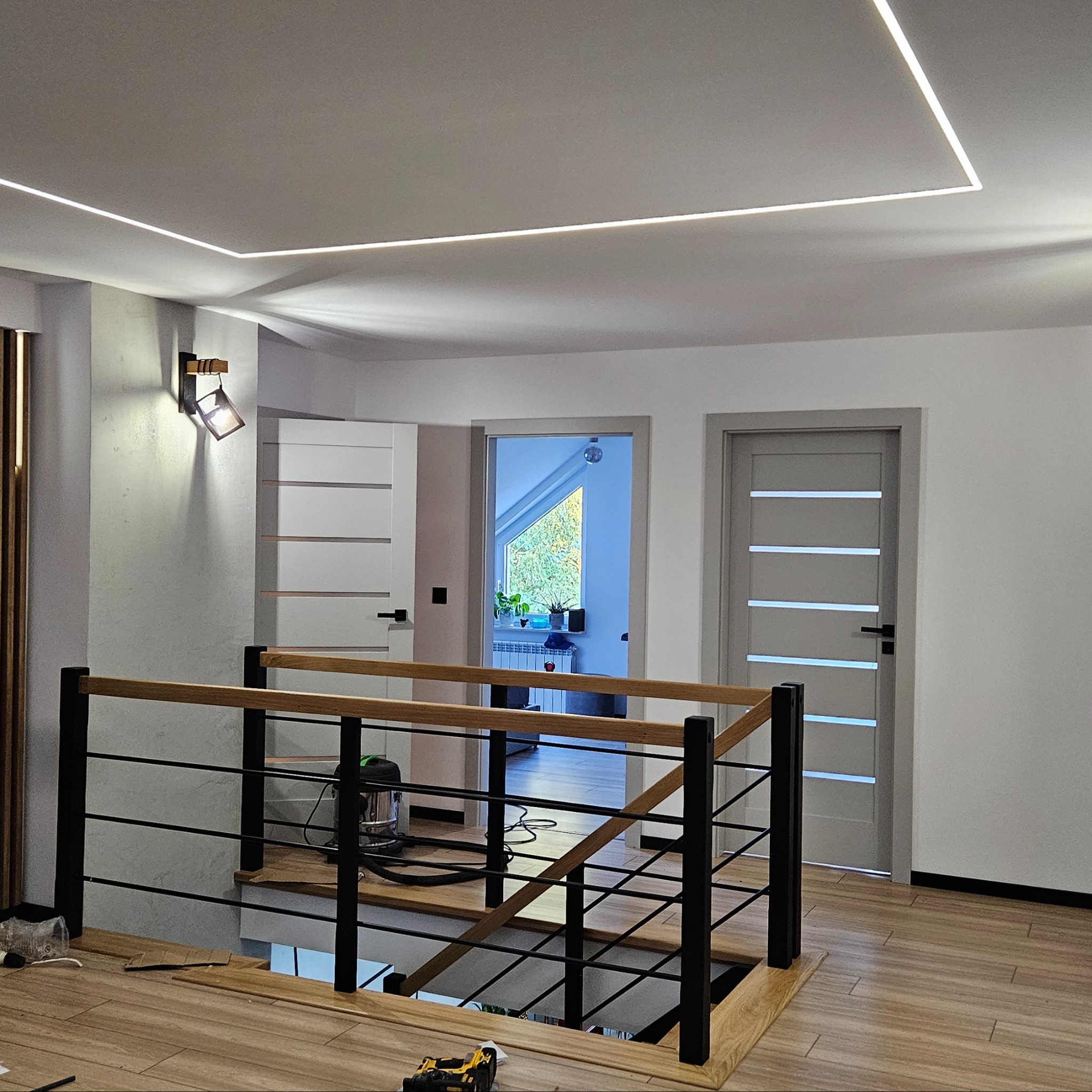 Nowoczesne oświetlenie LED w kształcie prostokąta na suficie w holu z drewnianą balustradą, widoczne drzwi i lampa ścienna. Częściowo widoczne narzędzia na podłodze.