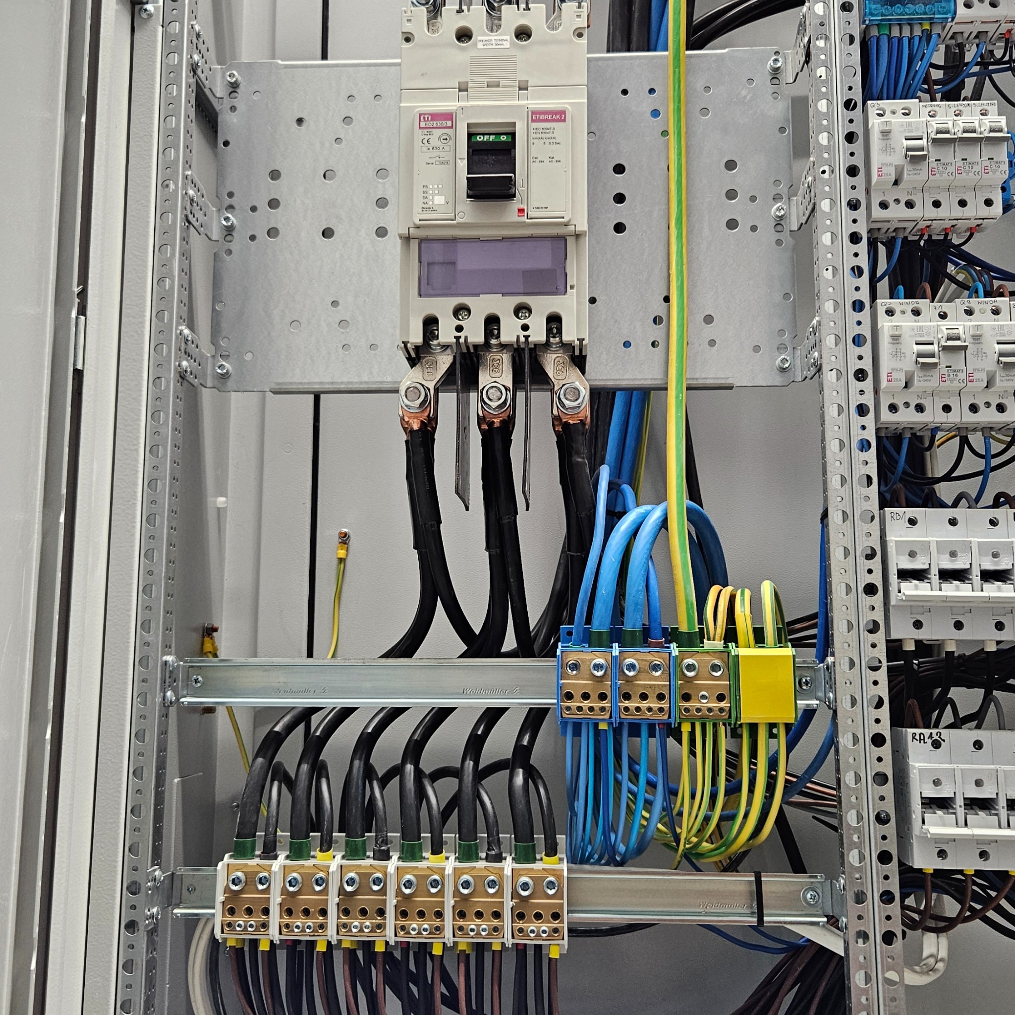 Rozdzielnica elektryczna z wyłącznikiem ETI ED2 630/3, licznymi przewodami w kolorach czarnym, niebieskim i żółto-zielonym, oraz listwami zaciskowymi Weidmuller, zamontowana na metalowej...