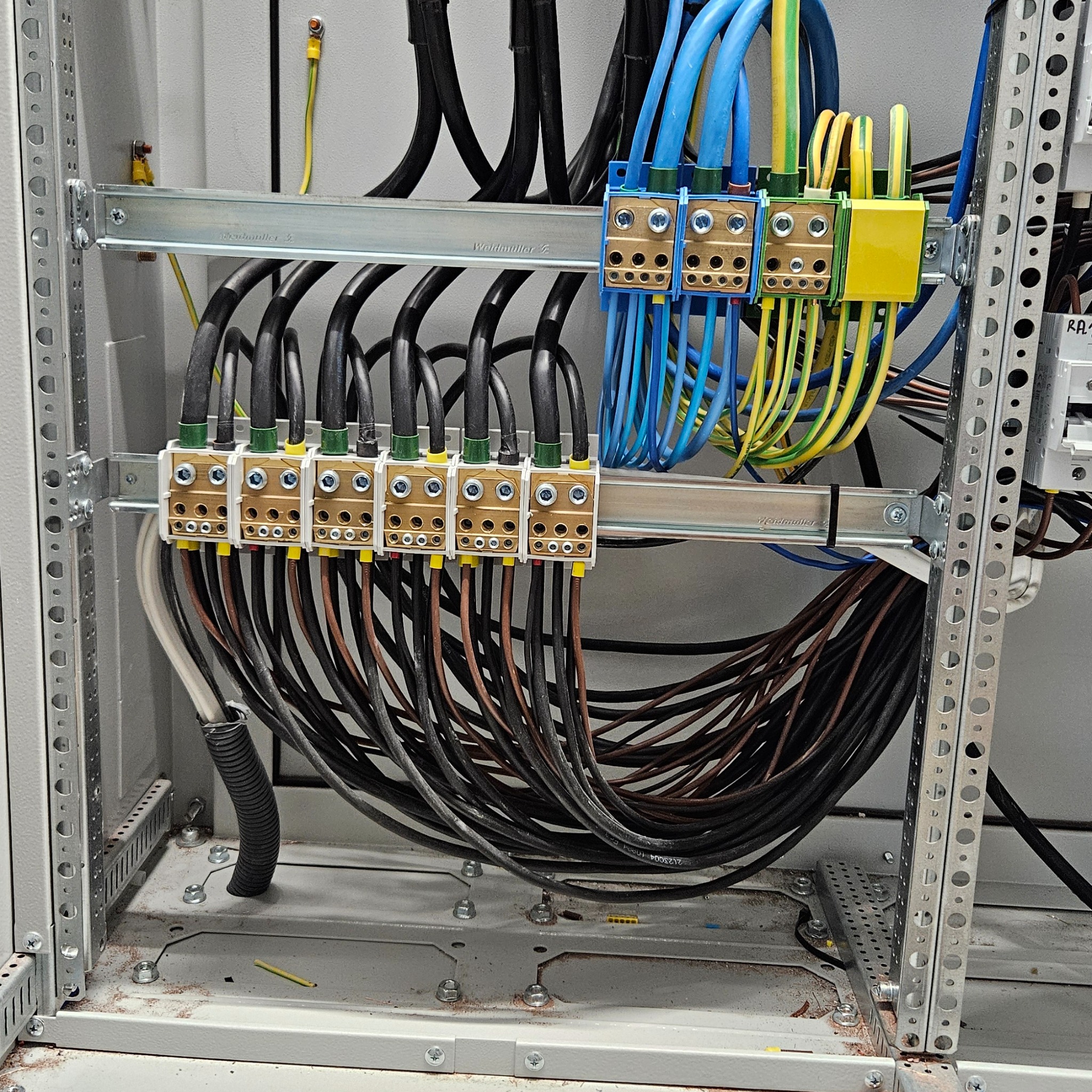 Wnętrze szafy sterowniczej z widocznymi listwami zaciskowymi, przewodami różnej grubości i koloru (czarne, niebieskie, żółto-zielone), oraz metalowymi szynami montażowymi.