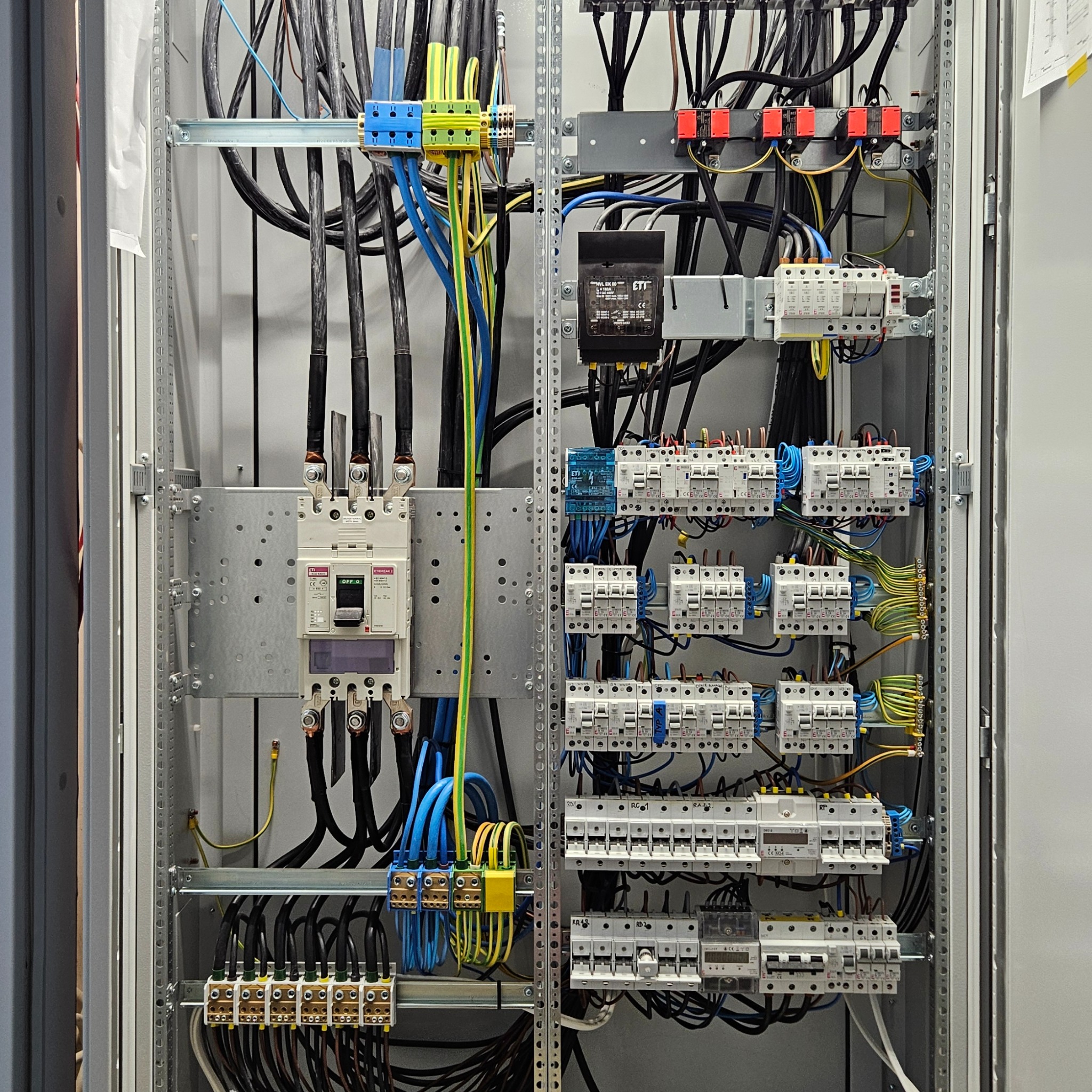 Rozdzielnia elektryczna w metalowej obudowie z widocznymi przewodami, bezpiecznikami i elementami sterującymi, uporządkowane okablowanie w kolorach czarnym, niebieskim i żółto-zielonym, widoczne...