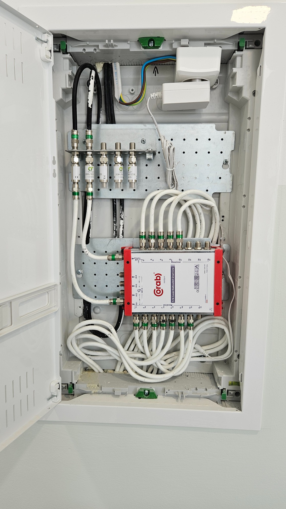 Rozdzielnica niskoprądowa z multiswitchem Corab i podłączonymi kablami koncentrycznymi, widoczne złącza F i przewody zasilające, zamontowana w białej obudowie w ścianie.