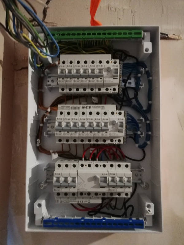 Otwarta skrzynka elektryczna z zamontowanymi bezpiecznikami, widoczne przewody w różnych kolorach, ściana w tle z widocznymi śladami po remoncie.