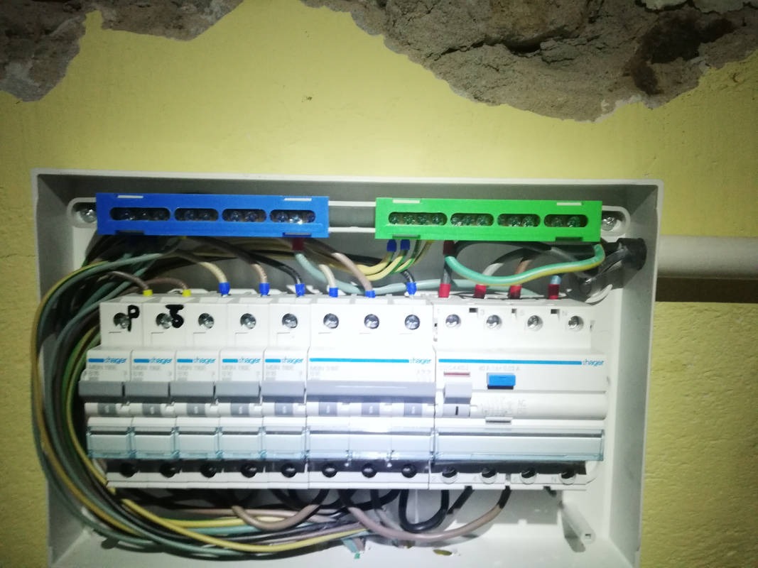 Rozdzielnica elektryczna Hager z podłączonymi przewodami, widoczne listwy zaciskowe w kolorze niebieskim i zielonym, montaż w żółtej ścianie z ubytkiem tynku.