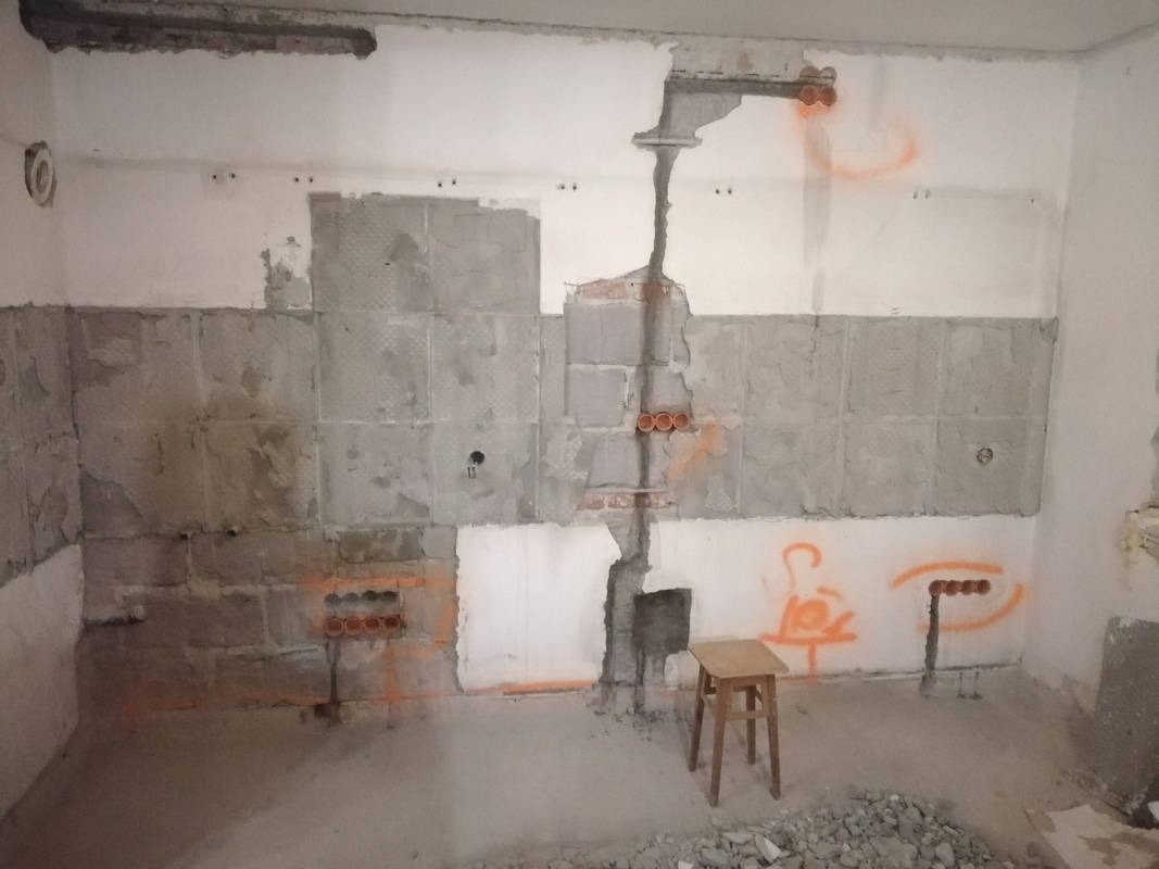 Fragment ściany w trakcie remontu z widocznymi bruzdami pod instalacje elektryczne, pomalowanymi na pomarańczowo otworami na puszki elektryczne i niewielkim drewnianym stołkiem na posadzce.