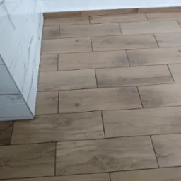 Ułożona podłoga z płytek imitujących drewno, widoczne fugi i fragment ściany wykończonej marmurowymi płytami oraz biały cokół.