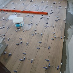 L..A Kora usługi remontowo budowlane - Układanie płytek podłogowych imitujących drewno, z użyciem klipsów poziomujących, widoczna poziomica i wiadro z klejem.