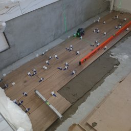 L..A Kora usługi remontowo budowlane - Układanie drewnopodobnych płytek podłogowych z użyciem poziomicy laserowej, poziomicy tradycyjnej i krzyżyków dystansowych; widoczny fragment ściany z ułożonymi białymi płytkami.