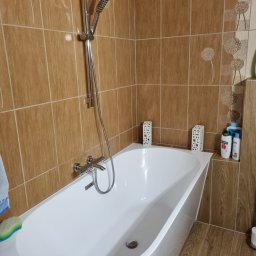 L..A Kora usługi remontowo budowlane - Biała wanna obudowana brązowymi płytkami z motywem dmuchawca, chromowana bateria i słuchawka prysznicowa, dekoracyjne ażurowe lampiony i kosmetyki na półce.