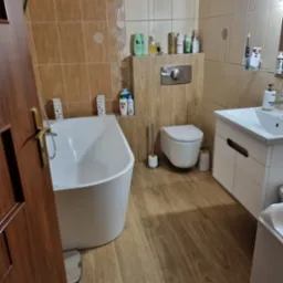 Wykończona łazienka w Wałbrzychu z brązowymi płytkami imitującymi drewno, białą wanną wolnostojącą, toaletą podwieszaną i umywalką z szafką, widok zza otwartych drzwi.