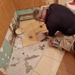 L..A Kora usługi remontowo budowlane - Mężczyzna w trakcie skuwania starych płytek podłogowych w łazience, widoczne fragmenty starej zaprawy i przygotowane nowe płytki.