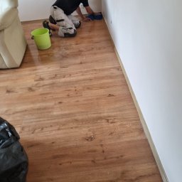 L..A Kora usługi remontowo budowlane - Mężczyzna na kolanach czyści ścianę w rogu pomieszczenia, widoczna podłoga z paneli, wiadro i fragment sofy.