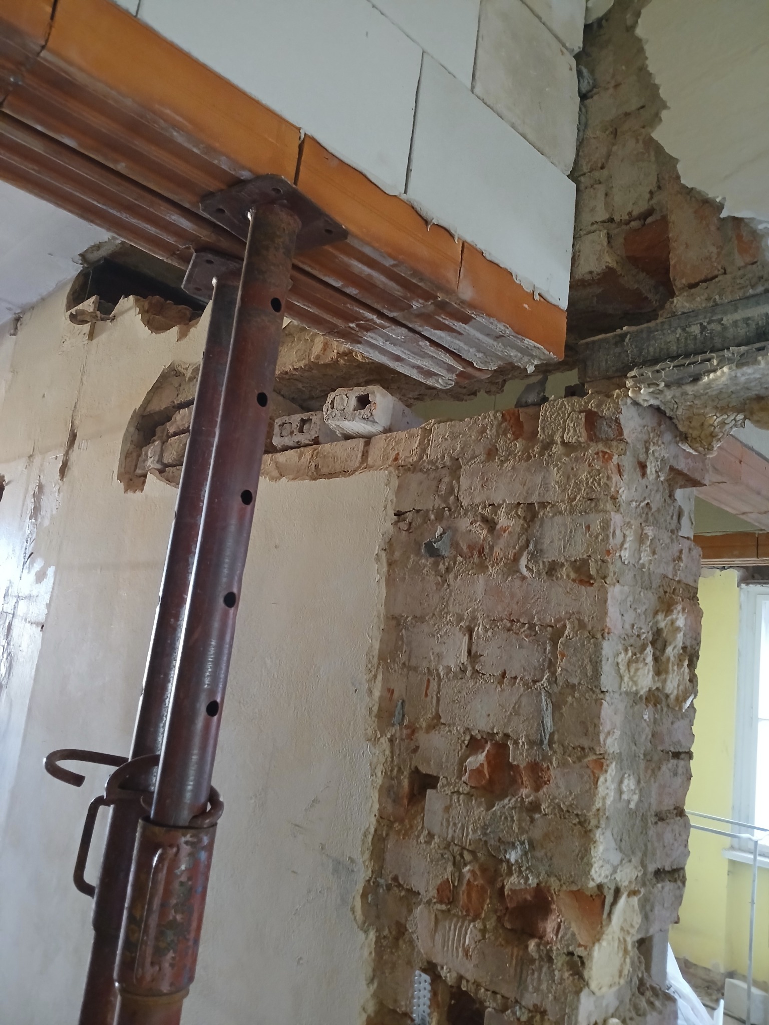 Podpory stropowe podtrzymujące drewniany element konstrukcyjny w trakcie prac remontowych, widoczne fragmenty muru z cegły i bloczków.